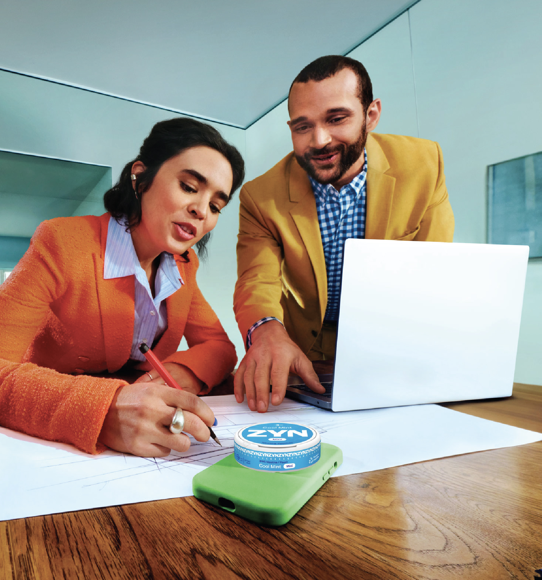A woman in an orange blazer sitting at a desk writing on paper in front of a laptop with a man in a yellow blazer leaning over. A white ZYN nicotine pouch container is placed on top of a phone in a green case on the table.